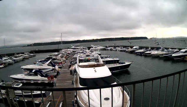 A marina filled with numerous boats docked along wooden piers, with a cloudy sky overhead and a distant shoreline in the background. The image appears to be captured from a webcam, showing the boats and water from an elevated perspective.