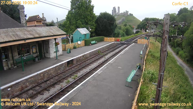 A small train station platform with benches, surrounded by green trees and flowers. A building with a stone facade and a corrugated roof is on the left, while a green shed and a yellow fence are further ahead. Two electrical poles with wires are on the right side, and railway tracks run through the scene towards a hill with ruins of an old castle in the distance. The sky is overcast. The image is marked with the date and time: 27-06-2025 at 19:00:24.
