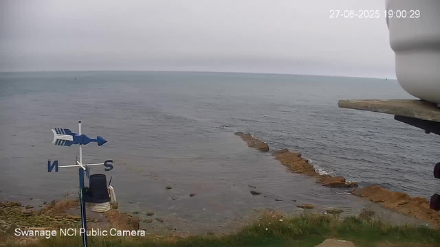 An overcast day at the seaside with a view of the water and a rocky shoreline extending into the sea. There is a weather vane on a pole in the foreground on the left, and part of a structure is visible on the right. The image is timestamped June 27, 2025, at 19:00:29.