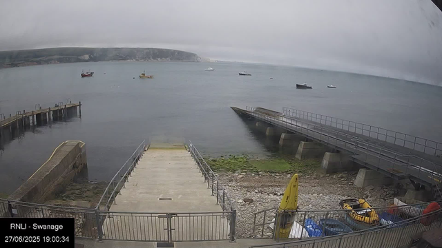 A view of a harbor on an overcast day with calm water. Several boats are anchored in the distance, and there are two boat ramps leading into the water. There is a small wooden dock on the left side, and a concrete slipway with a metal railing on the right side. In the foreground, there are some yellow safety equipment and a yellow inflatable boat stored near the railing. The shoreline is rocky, and a grassy patch is visible near the bottom right corner. The background features a distant coastline with cliffs and low clouds covering the sky.