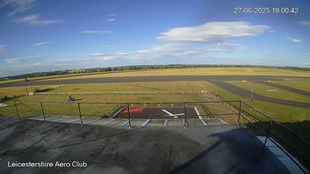 A wide view of an airfield with a runway extending across the horizon under a partly cloudy sky. In the foreground, there is a helipad with marked landing zones and a small fence surrounding it. A bench and a windsock are visible nearby. The bottom corner shows the text "Leicestershire Aero Club," and the top right corner displays the date and time as "27-06-2025 19:00:42."
