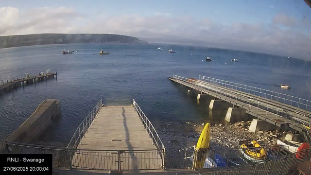 A view of a harbor with a concrete pier extending into calm water, boats anchored in the distance, and a distant coastline with cliffs under a partly cloudy sky. There is a yellow kayak and some other small boats visible near the shore. The timestamp in the bottom left corner reads "27/06/2025 20:00:04," and the location is noted as "RNLI - Swanage."