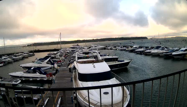 A marina filled with numerous boats and yachts docked along wooden piers, with a large white yacht prominently positioned in the center. The sky is cloudy with a mix of light and dark clouds, and the water appears calm. In the background, there is a distant shoreline with some land visible.
