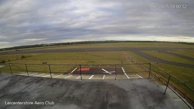 A view from a webcam at Leicestershire Aero Club shows an empty apron area with a nearby runway extending into a flat, open field. The sky is overcast with gray clouds. There is a small safety grid painted on the ground in the foreground, and a windsock on a pole to the left. In the distance, grassy fields and trees stretch toward the horizon. The timestamp on the image reads "27-06-2025 20:00:52."