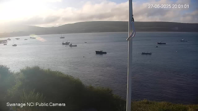 A view of a calm body of water with several boats floating on the surface. In the background, there are hills under a cloudy sky. On the right side, part of a vertical pole or mast is visible. The timestamp in the upper right corner shows the date and time as 27-06-2025 20:01:01. The bottom left corner has the text "Swanage NCI Public Camera."