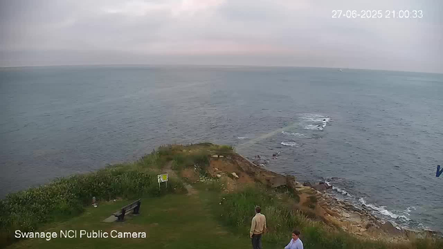 A coastline scene with a grassy area in the foreground, where two people are standing and talking. One person is facing away from the camera, wearing a beige jacket, while the other is partially visible, wearing a light-colored shirt. There is a bench on the grass near some bushes. The rocky shoreline extends into the water, which appears calm with gentle waves. The sky is cloudy with a light pink hue near the horizon. A small boat is visible in the distance on the water. The timestamp in the top right corner shows "27-06-2025 21:00:33," and the bottom left indicates the camera is a "Swanage NCI Public Camera."