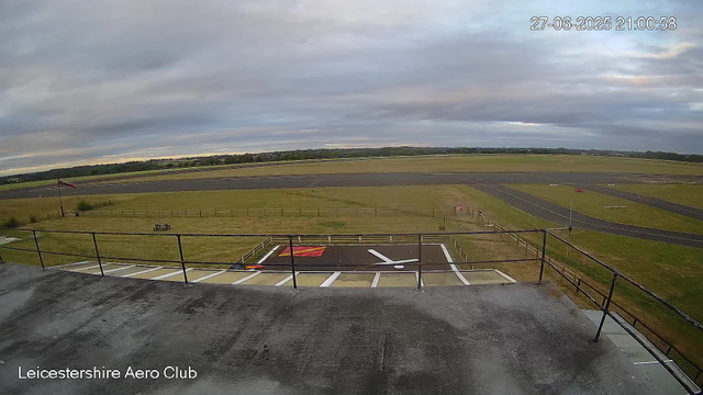 A view from a webcam at Leicesterhire Aero Club showing an airfield with a grass runway and a paved taxiway. The sky is overcast with gray clouds, and the surrounding landscape is flat with fields and distant trees. There is a windsock on a pole near the runway, and a marked helicopter landing pad in the foreground. The timestamp on the image is 27-06-2025 at 21:00:58.