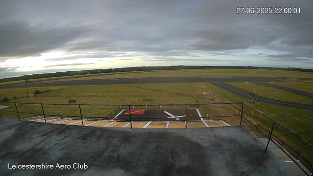 An outdoor view from a webcam at Leicestershire Aero Club shows a cloudy sky over a runway and surrounding grassy area. In the foreground, there is a helipad with markings, a railing, and a small platform. A windsock is visible on the left side, and the vast open fields with patches of runway extend into the distance under a moody sky. The timestamp reads 27-06-2025 22:00:01.