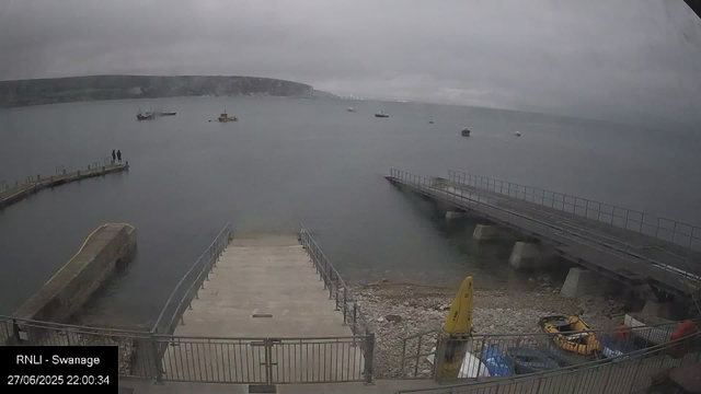 A view of a cloudy, overcast day at the harbor in Swansea. There are two people standing on a small pier that extends into calm water. Several boats are anchored in the distance. In the foreground, there is a concrete slipway with a railing, a yellow warning sign, and some parked vehicles. The background features a coastline with cliffs or hills shrouded in mist. The bottom left corner displays a timestamp and the label "RNLI - Swansea."