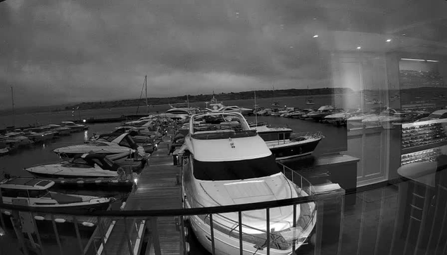 A black-and-white photograph of a marina taken from inside a building through a window. The image shows several boats and yachts docked at the pier, with calm water surrounding them. In the background, there is an open sky with some clouds and land visible on the horizon. Inside the room, faint reflections of furniture and the interior can be seen overlaying the view.