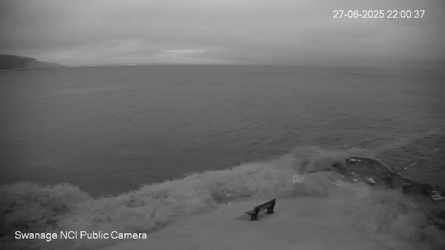 A black and white image from a webcam shows a coastal scene with an empty bench near the water's edge. Waves are gently crashing onto the shore, and a small boat or dock is partially visible on the right side. The horizon line separates the water from a cloudy sky in the background. The image has a timestamp of June 27, 2025, at 10:00 PM, and a label indicating it is from the Swansea NCI Public Camera.