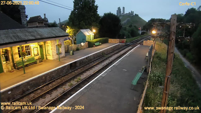 A small train station platform is shown with a building on the left and a row of green benches. There are flower pots hanging from the building, and a "Way Out" sign near the entrance. The platform is empty and illuminated by several warm yellow lights. To the right, there are railway tracks running parallel to the platform, with an overhead electric line supported by wooden poles. In the background, a hill with the ruins of an ancient castle or fortress is visible, surrounded by greenery and trees. The scene appears to be early evening or dusk, with a cloudy sky. The timestamp indicates the time as 22:00:48 on June 27, 2025.