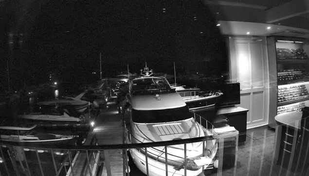 A black-and-white image taken from a webcam shows the interior of a yacht club or marina at night. Several yachts and boats are docked outside, with some illuminated by small lights. Inside, a section of the room displays a bar or display shelf filled with bottles, and there's a curved glass railing in the foreground. The scene is dark, with some ambient indoor lighting visible.
