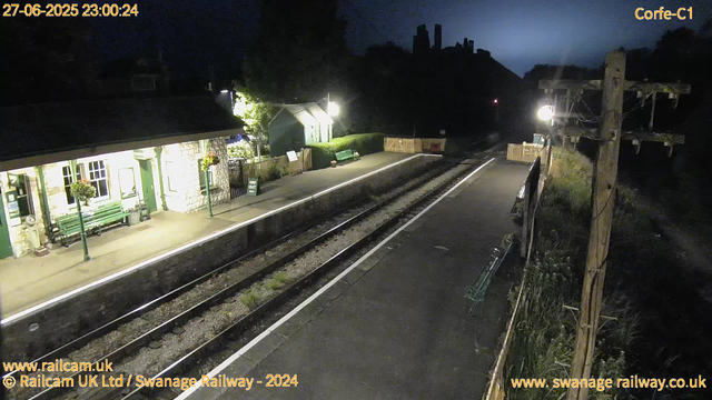 A small train station platform at night, illuminated by bright lights. The platform has green benches, flower baskets, and a small shelter. There is a sign saying "WAY OUT" and a trash bin next to the shelter. The railway tracks run through the middle of the scene, with a path on the right side. In the background, there are dark hills with some industrial structures on top, silhouetted against the night sky. On the right side, there are wooden utility poles and some fencing and vegetation. The image includes a timestamp and website information at the top and bottom borders.