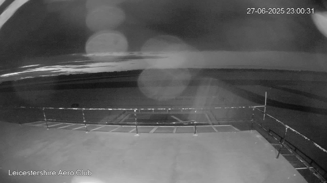 A black and white image from a webcam showing an empty parking lot at night. The parking lot is partially lit, with a few marked parking spaces visible. In the background, there is a body of water with some reflections and distant lights. The image has a timestamp in the upper right corner showing the date "27-06-2025" and the time "23:00:31". The lower left corner displays the text "Leicestershire Aero Club".