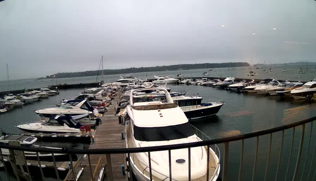 A marina filled with numerous boats and yachts docked along wooden piers. The water surrounds the boats, with a distant shoreline and a cloudy sky overhead.