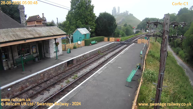 A small railway station platform with a stone building on the left side, featuring a sloped roof and a chimney. There are green benches along the platform and a small green shed behind a wooden fence. Railway tracks run through the center, with a signal in the distance. An old wooden electric pole with wires extends along the right side, next to a grassy area with wildflowers and a narrow footpath. In the background, a hill with ruins or castle remains is visible, partially obscured by light fog or mist. The timestamp indicates it is early morning at 5:00 AM.