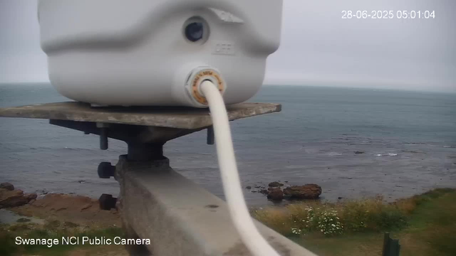 A close-up of a white device mounted on a weathered wooden and metal stand overlooking a coastline with rocks in the water and some green vegetation in the foreground. The device appears to be a camera or sensor, with a cable connected to it. The background shows an overcast sky and the sea stretching into the distance. The timestamp in the upper right corner indicates the date and time: 28-06-2025, 05:01:04. The text at the bottom left reads "Swanage NCI Public Camera."