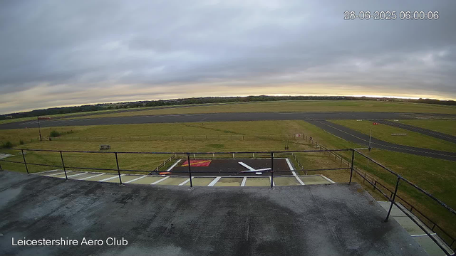 A wide view of an airfield runway under a cloudy sky. In the foreground, there is a small tarmac area with white parking lines, a red cover, and a black arrow painted. A flagpole with a red and white flag, a bench, and a safety railing are also visible. The background features an open grassy area with a runway extending into the distance. The timestamp in the upper right corner shows the date and time as 28-06-2025 06:00:06. The bottom left corner indicates "Leicestershire Aero Club."