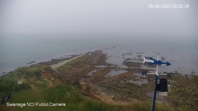 A wide view of a rocky shoreline and tidal area at low tide, with a narrow pier extending into the water. The sky is overcast and foggy, creating a gray atmosphere. There is a weather vane and an anemometer mounted on a pole in the foreground on the right side. In the lower left corner, a small green grassy area with a bench and a sign. The image has a timestamp in the upper right corner reading "28-06-2025 06:00:38," and a label in the bottom left corner that says "Swanage NCI Public Camera."