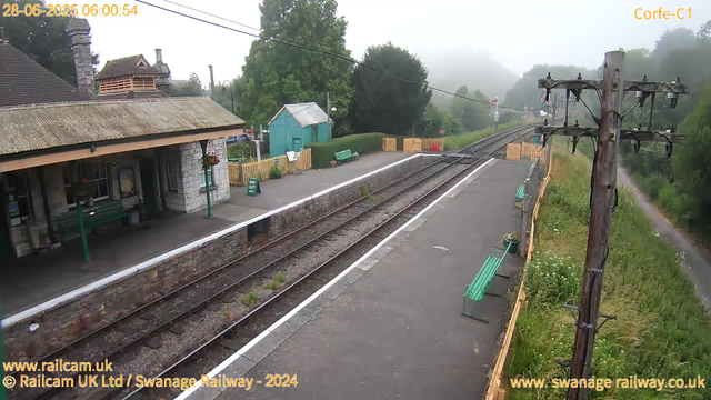 A view of a small railway station platform on a foggy morning. There are green benches along the platform, a small blue shed, and a yellow fence bordering the platform. An electrical pole with wires runs along the right side of the image. In the background, there are trees and a mist-covered landscape. The station building has a stone exterior with a window and flower baskets hanging on the wall. The scene is calm, with no trains or people visible at the moment.