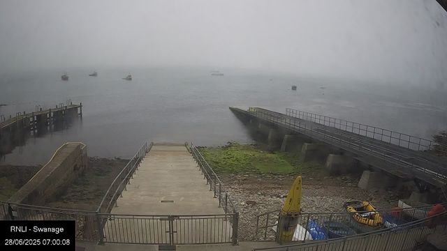 A foggy seaside scene showing a small concrete pier extending into calm water. There are several boats in the distance, mostly obscured by the fog. A sloped walkway with railings leads down to the water on the right, alongside a ramp that appears to be for moving boats or equipment. In the foreground, there's a fenced area with some yellow equipment, a yellow caution sign, and a few plastic containers. The timestamp in the lower left corner reads "28/06/2025 07:00:08" and the location is noted as "RNLI - Swanage."