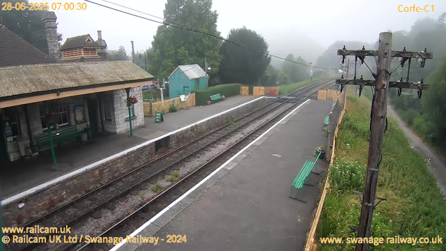 A small railway station platform is visible with green benches and a "Way Out" sign near the building. The building has stone walls, a tiled roof, and hanging flower baskets. Railway tracks run through the scene, surrounded by greenery and trees. A tall wooden telegraph pole with multiple wires is nearby. The sky and distant background appear foggy, and the overall atmosphere suggests early morning with limited visibility. The date and time in the corner indicate it is 7:00 AM on June 28, 2025.