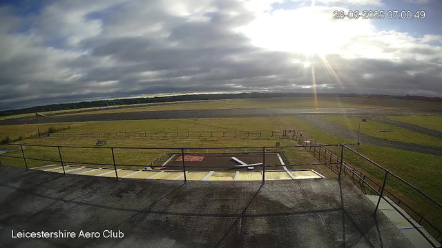A view of an outdoor airfield taken from a webcam at Leicesterhire Aero Club. The image shows a grassy area with a small, fenced boundary, a paved walkway, and an empty runway stretching into the distance. The sky is partly cloudy, with the sun shining brightly from the upper right, creating lens flare. There are some distant trees on the horizon. The timestamp in the upper right corner reads 28-06-2025 07:00:49.