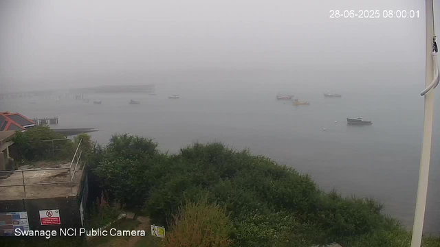 A foggy scene over a body of water with several boats floating on the surface. In the foreground, there are green bushes and a small building with a red roof. There are some signs and a railing near the building. The image has a timestamp in the upper right corner showing the date as June 28, 2025, and the time as 8:00:01 AM. The bottom left corner indicates the footage is from the Swannage NCI Public Camera.