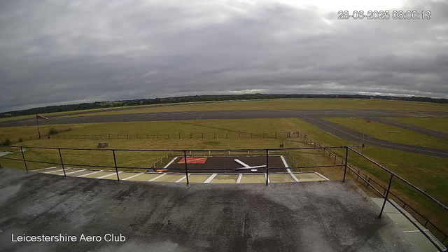 A view from a webcam at Leicestershire Aero Club shows an overcast sky with dark clouds. In the foreground, there is a fenced area with a small red and black helipad or parking space, and a railing along a balcony or observation deck. Beyond, there is a grassy field with a windsock on a pole, and a paved runway extending into the distance. The landscape is flat with some trees and open land in the horizon. The timestamp on the image is 28th June 2025 at 8:00 AM.