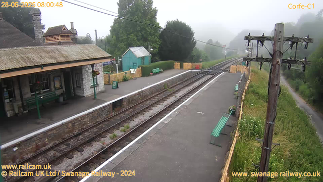 A small train station platform on a foggy morning. There are two green benches along the platform, a small blue building, and wooden fencing. An electrical pole with overhead wires is visible on the right side. The railway tracks run alongside the platform, curving into a fog-covered landscape with trees in the background. A building with a tiled roof and chimney is partially visible on the left. The scene includes some greenery and a narrow path running parallel to the tracks on the right side. The timestamp reads "28-06-2025 08:00:48."