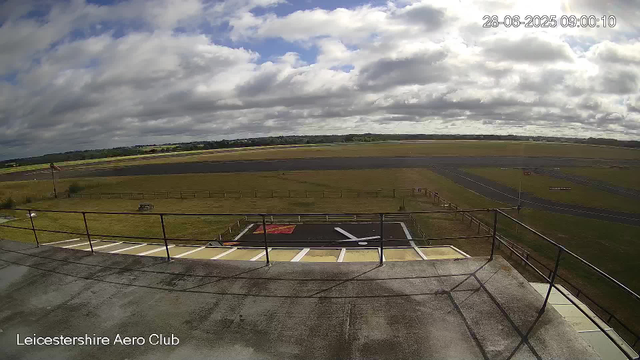 A view from a webcam at Leicesterhire Aero Club showing an open sky with scattered clouds, a runway in the distance, and a small paved area with a railing in the foreground. The timestamp indicates it was taken on June 28, 2025, at 09:00:10.