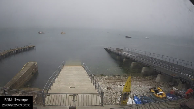 A foggy harbor scene with several boats floating on calm water. There are two dock structures extending into the water, one on the left and one on the right, with railings. In the foreground, there is a concrete and gravel area with yellow equipment, possibly life-saving devices, and a small boat or kayak. The date and time displayed are June 28, 2025, at 9:00:38 AM.
