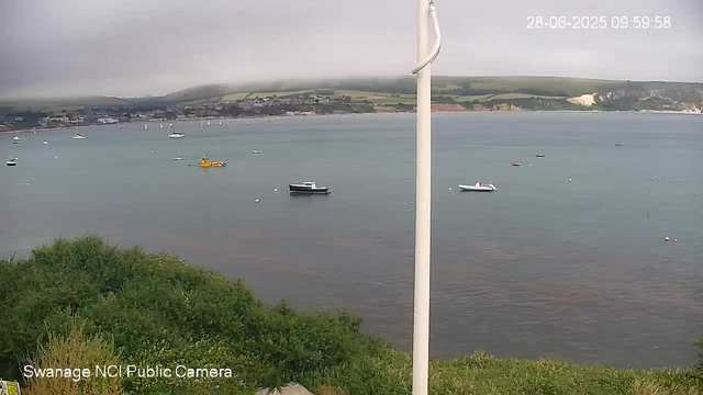 A view of a calm harbor with several boats floating on the water. The shoreline with green foliage is in the foreground, and a distant town and hills are visible in the background under a cloudy sky. The image is timestamped "28-06-2025 09:59:58" in the upper right corner.