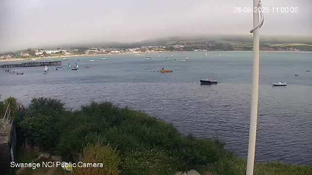 A view of a harbor with several sailboats and small boats floating on calm water. In the background, there is a shoreline with buildings and a pier extending into the water. The sky is overcast and appears misty or foggy, with low-hanging clouds covering the distant hills. The timestamp on the top right corner indicates the date as August 28, 2025, and the time as 11:00:06. In the lower left corner, there is a label that reads "Swanage NCI Public Camera."