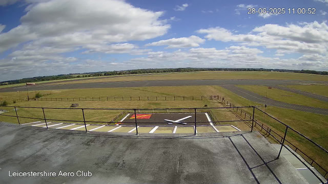 A wide view of the Leicesterhire Aero Club airfield under a partly cloudy sky. In the foreground, there is a helipad marked with a white 'H' and surrounded by white lines, with a red and yellow boundary on the ground. A metal railing runs along the edge of the viewing platform. Beyond the helipad, there is a grassy area with a small picnic table, and a windsock on a pole indicating the wind direction. In the background, there are runways and taxiways extending across the field, with some distant trees on the horizon. The timestamp indicates the date and time as 28-06-2025, 11:00:52.