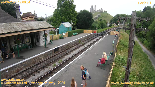 A small train station platform with a few people waiting. There are green benches, a wooden fence, and a small green building. In the background, there is a hill with a castle ruins on top. To the right, a wooden utility pole with wires extends along a narrow grass path. The sky is slightly overcast. The timestamp shows "28-06-2025 12:00:04".