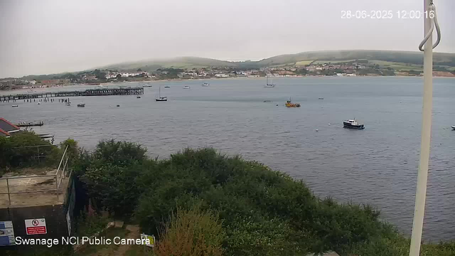 A view of a harbor with several small boats floating on calm water. In the background, there are hills and a town with buildings along the shoreline. In the foreground, there are some green bushes and part of a construction area with a sign, and a vertical pole on the right side of the image. The timestamp in the upper right corner indicates the date and time as June 28, 2025, at 12:00:16. The bottom left corner has the label "Swanage NCI Public Camera."