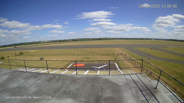 A view from a webcam at Leicestershire Aero Club shows an empty outdoor runway and taxiway under a blue sky with scattered clouds. In the foreground, there's a fenced observation deck with a small painted landing area on the ground. The landscape beyond features grassy fields and the horizon in the distance. The timestamp on the image reads 28-06-2025 12:00:56.