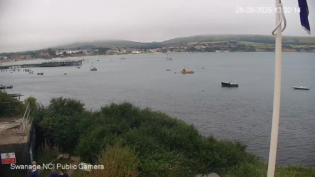 A view of a harbor with several boats floating on calm water, a pier extending into the harbor on the left, green hills with scattered buildings in the background, and a cloudy sky overhead. A white flagpole with a partially visible flag is in the foreground on the right. The timestamp shows the date and time as 28-06-2025 at 13:00:14.