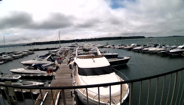 A marina filled with numerous boats and yachts docked along wooden piers extends into calm water under an overcast sky, with a distant shoreline visible on the horizon.