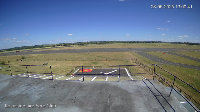 A wide view of an airfield with a clear blue sky and scattered clouds. In the foreground, there is a helipad with a white "H" marking and a red square nearby, surrounded by a black safety railing. A windsock is visible to the left, indicating wind direction. The runway extends into the distance with grassy areas on either side, and some distant trees and structures are visible on the horizon. The timestamp in the upper right corner reads "28-06-2025 13:00:41," and the bottom left corner displays "Leicestershire Aero Club."