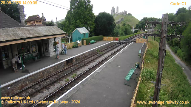 A small railway station platform with a stone building on the left side, where a few people are standing and sitting. There are green benches on the platform, a fenced area with a small turquoise shed, and a row of flowers in pots along the fence. The railway tracks run through the middle of the image, stretching into the distance with lush green trees and hills surrounding the station. In the background, on a hill, there are ruins of an old castle or stone structure. An old wooden telephone pole with wires is on the right side, and a narrow footpath runs alongside the tracks. The sky is partly cloudy with some sunlight.