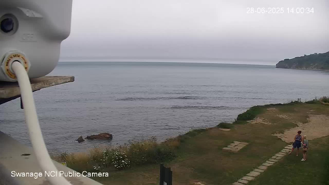 A view of a cloudy seaside landscape, with a calm ocean stretching to the horizon and a distant headland on the right side. In the foreground, there is a grassy area with a stone pathway, and two people standing and talking near the water's edge. There is also a partial view of a white camera on the left side of the image. The timestamp in the upper right corner reads "28-06-2025 14:00:34".
