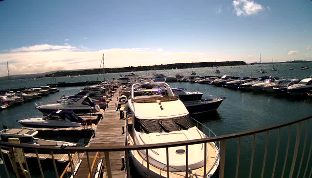 A marina filled with numerous boats and yachts docked along wooden piers extending into calm water. The sky is partly cloudy with patches of blue, and distant land or shoreline is visible across the water.
