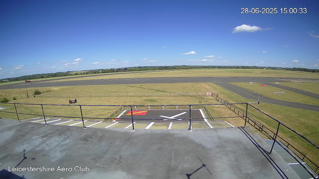 A clear view from a webcam at Leicestershire Aero Club shows an empty tarmac runway extending into a grassy airfield under a bright blue sky with scattered white clouds. In the foreground, there is a small helipad marked with a white "H" and a red square, surrounded by a white and orange border. A black bench with a wooden seat faces the airfield. A few small signs and barriers are situated around the helipad area. The wide open space stretches into distant fields and hills, creating a peaceful rural scene. The timestamp in the top right corner indicates the date as June 28, 2025, and the time as 15:00:33.