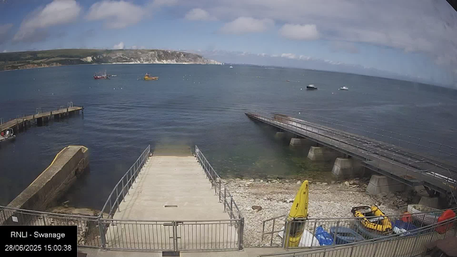 A view of a harbor with a concrete dock area in the foreground. There are two small boat ramps ledking into the water, and a yellow kayak and related gear are visible near the right side. Several small boats are moored in the water, with a few more boats in the distance. In the background, there are cliffs and hills along the coastline, under a partly cloudy sky. The image includes a timestamp and the label "RNLI - Swanage" at the bottom left.