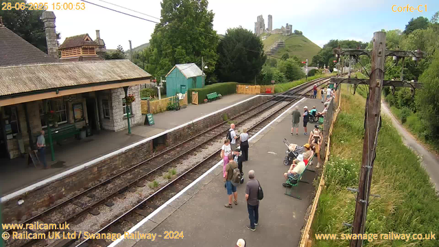 A small train station platform with several people waiting, some sitting on benches and others standing or walking. There is a stone building with a tiled roof on the left side and a few colorful buildings and trees behind it. A row of train tracks runs through the middle, with greenery and trees along the sides. In the background, a hill with the ruins of a castle is visible. Overhead, there are utility poles with wires. The scene appears to be during daytime on a partly cloudy day.