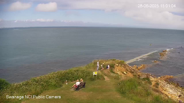 A view of a coastline with a grassy area and a few people sitting on a bench and standing near the rocks. The ocean stretches out to the horizon under a partly cloudy sky. There are some boats in the water, and a breakwater extends into the sea on the right side. The timestamp in the top right corner shows the date and time as 28-06-2025 15:01:04.