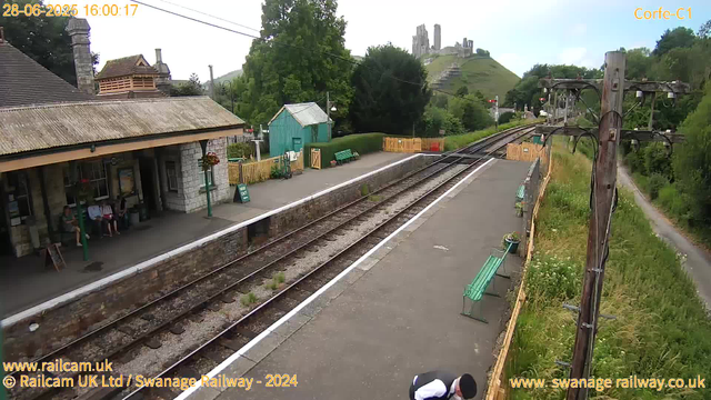 A small railway station platform with a building on the left, where three people are seated under a covered area. There are green benches along the platform, a small green shed, and a yellow fence at the far end. A few plants are in pots along the platform edge. To the right, there are electrical poles with wires. In the background, there is a hillside with an ancient castle ruins on top, surrounded by trees and greenery. The sky is partly cloudy.